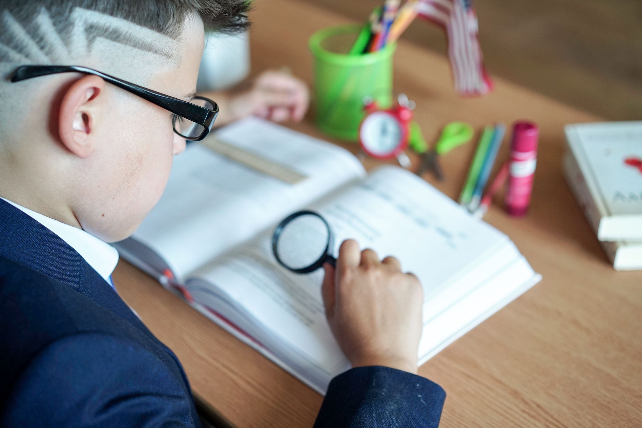 A young boy focuses on his book using a magnifying glass at a study desk.