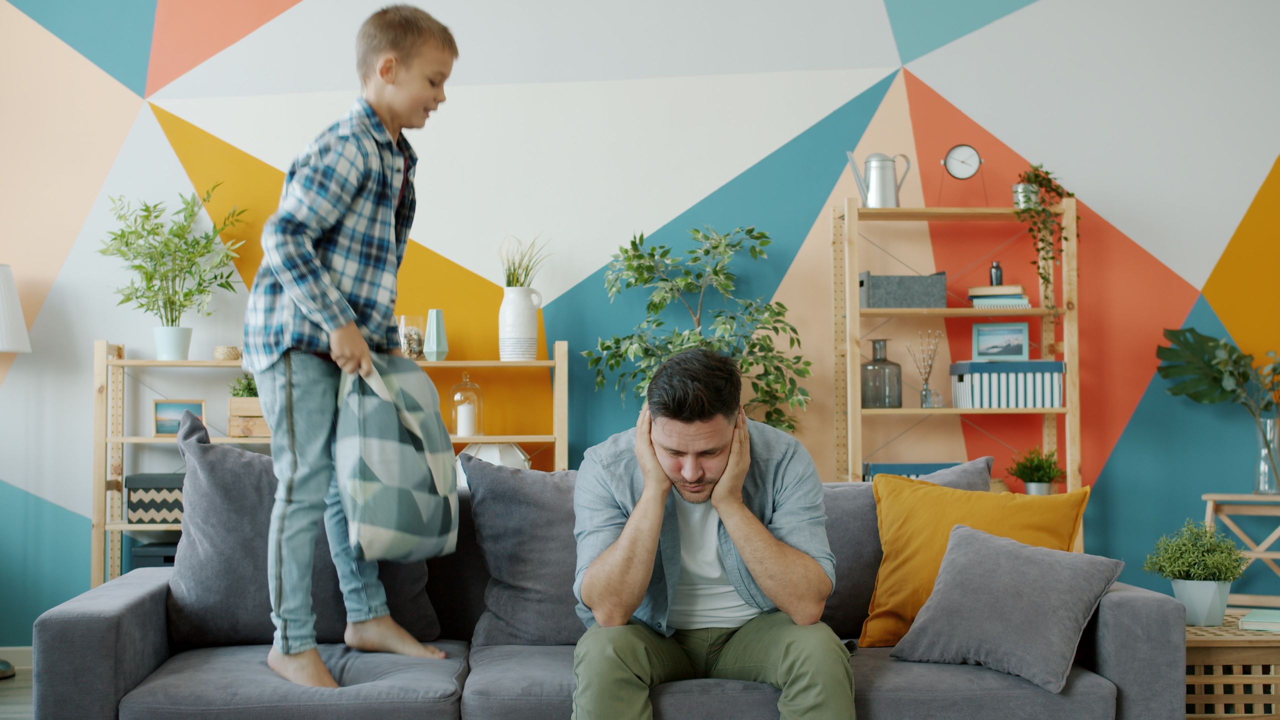 A young boy playfully jumps on the couch beside his father holding a pillow, creating a lively home atmosphere.