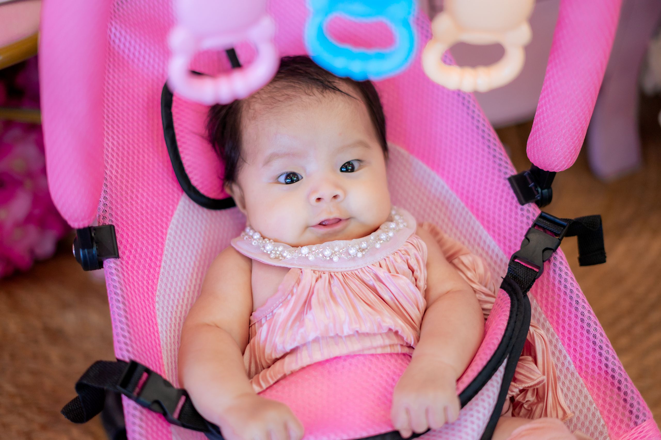 Adorable baby lying in a pink bouncer seat, dressed in a peach outfit indoors.