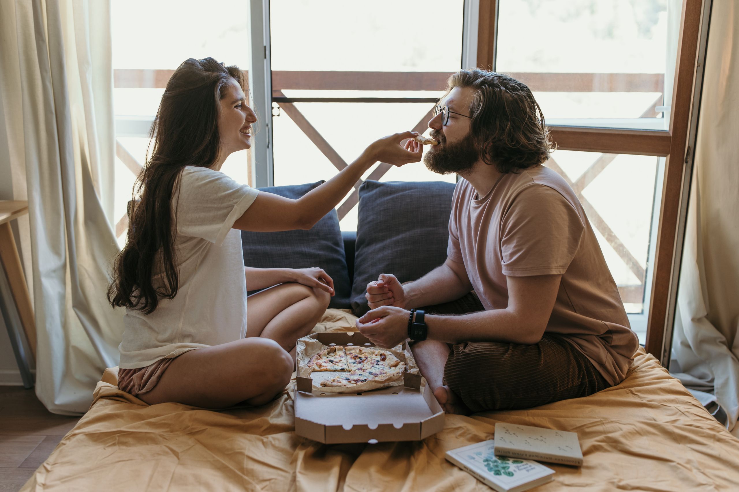 Couple enjoying a cozy moment sharing pizza on a bed by a window. Perfect for lifestyle and relationship themes.