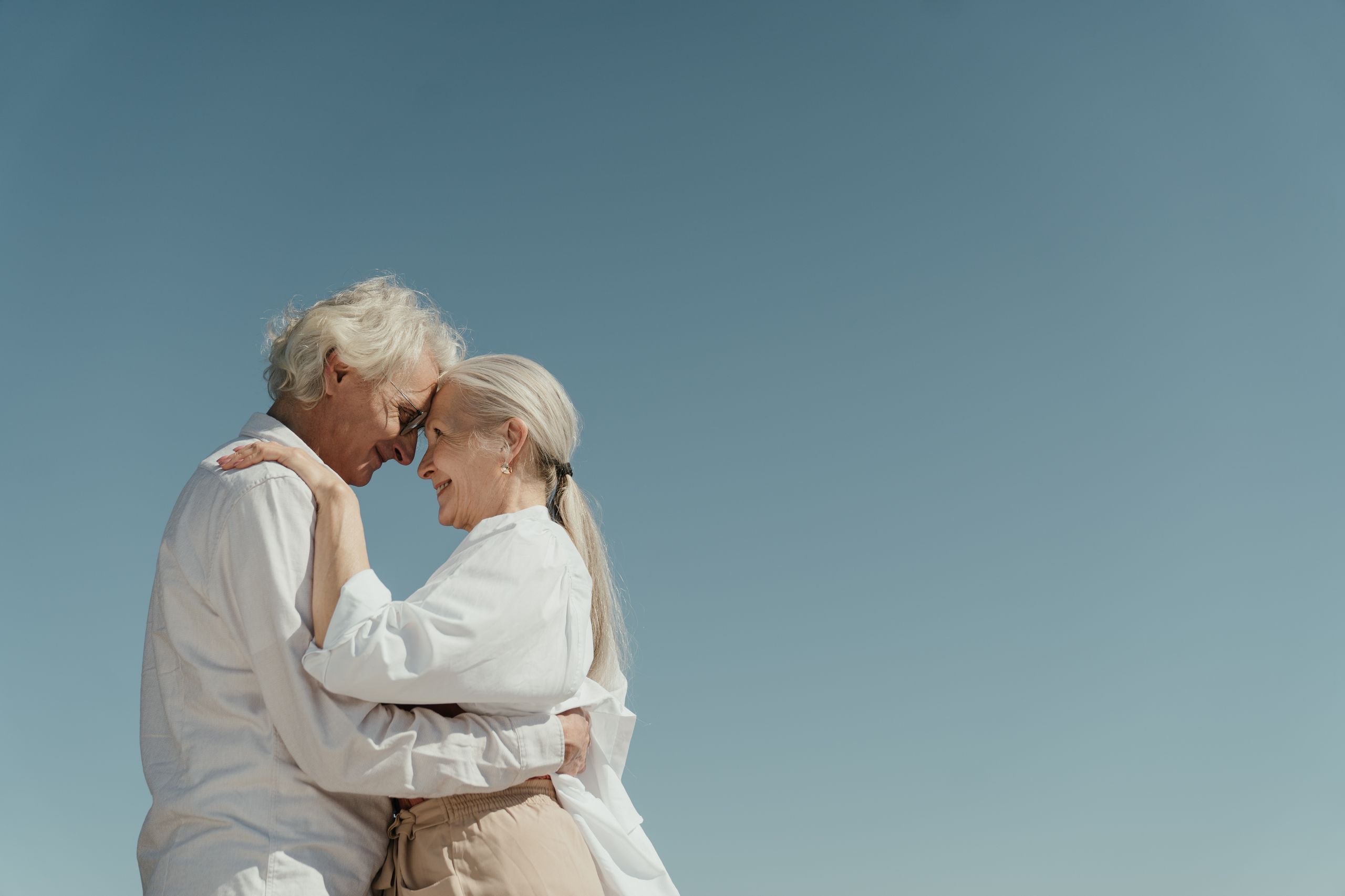 Senior couple embracing outdoors, depicting love and happiness against a clear blue sky.